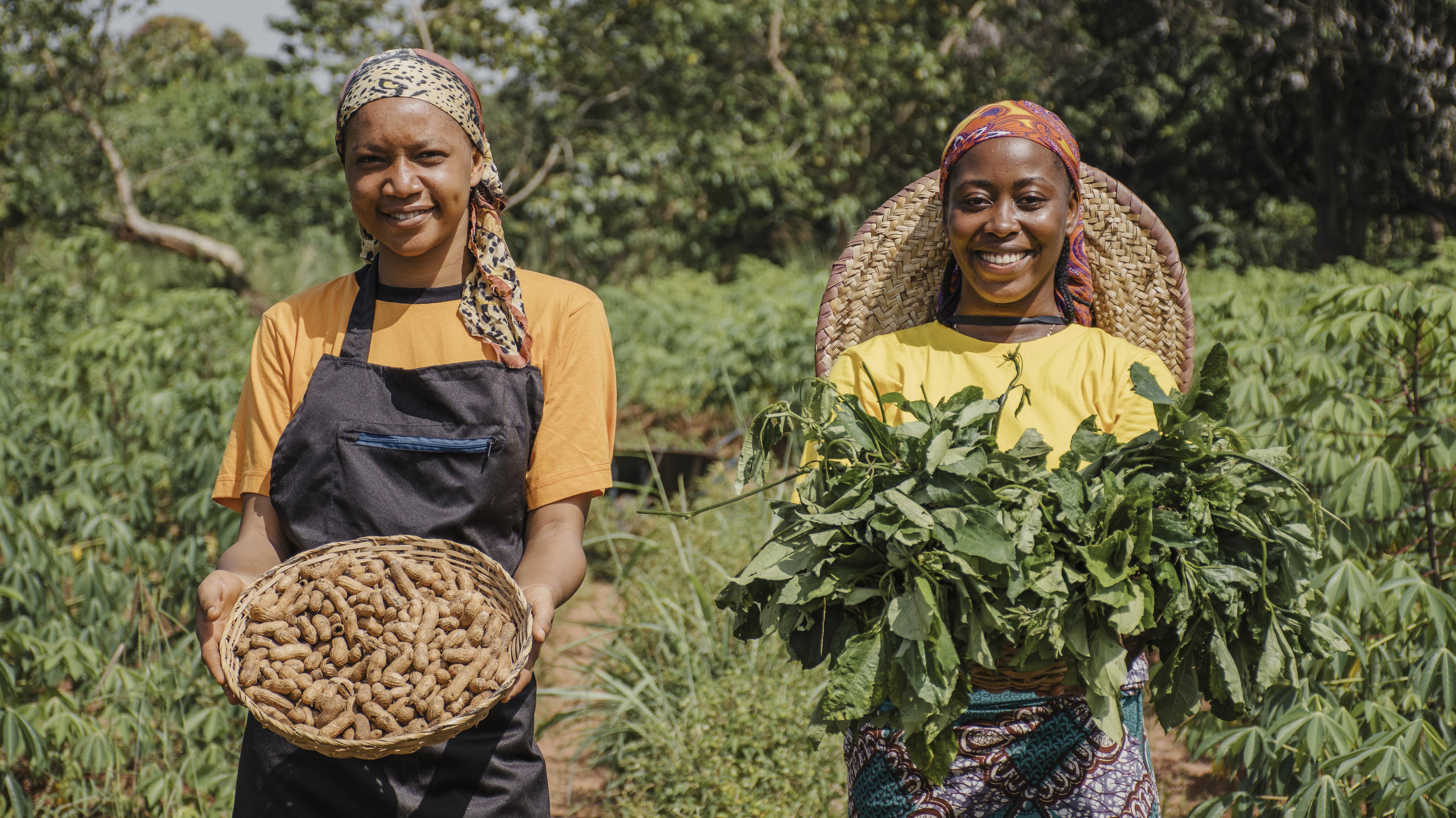 Two women farmers posing proudly with baskets of peanuts and fresh greens in a lush field